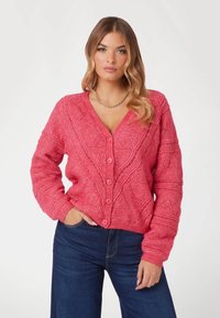 Cardigan rosa lavorato a maglia con un motivo testurizzato, dotato di scollo a V, chiusura con bottoni e polsini a costine. Abbinato a jeans blu scuro.