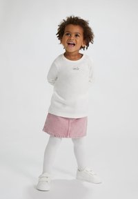 Witte longsleeve shirt met de tekst "smile", gecombineerd met een roze dennen rok, witte maillots en witte klittenbandsneakers.