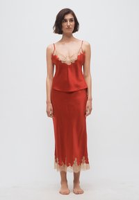 Vrouw die een rode satijnen slipjurk draagt met beige kanten bies bij de halslijn en zoom, blootsvoets staand tegen een effen witte achtergrond.