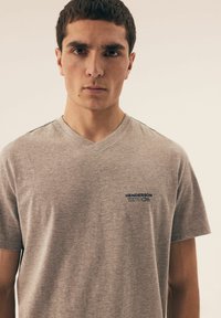 Szara bawełniana koszulka z dekoltem w serek i krótkim rękawem. Posiada małe haftowane logo w kolorze niebieskim na lewej stronie klatki piersiowej. Gładka faktura.