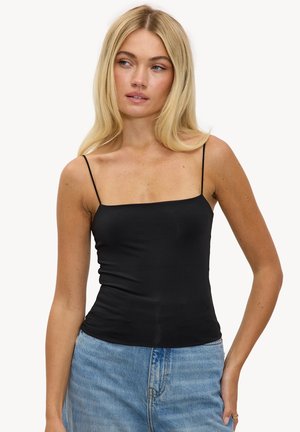 Femme blonde portant un haut noir à bretelles fines et un jean taille haute bleu clair, regardant légèrement vers la droite avec une main dans la poche.