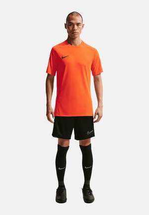 Mężczyzna stojący, ubrany w pomarańczowy krótki rękaw Nike, czarne spodenki Nike, czarne skarpety do kolan i czarne korki piłkarskie.
