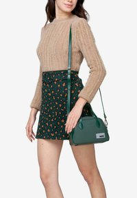 Groene leren handtas met gestructureerde vorm en korte handvatten, gedragen bij een beige fluffy trui en groene luipaardprint rok.