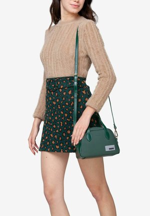 Sac à main en cuir vert avec une forme structurée et des poignées courtes, porté avec un pull en laine beige duveteux et une jupe verte à imprimé léopard.