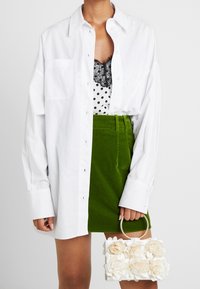 Chemise blanche oversize, haut en dentelle à pois noirs, jupe en velours côtelé verte, sac à main blanc avec ornements floraux, anse en anneau argenté.