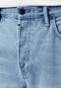 Svetlo modre kavbojke iz jeansa z zapiranjem na gumb, sprednjo zadrgo in všitimi žepi. Imajo rahlo teksturirano tkanino in klasičen kroj.