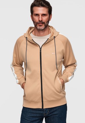 Homme barbu portant un sweat à capuche beige zippé avec cordons noirs et manches rayées blanches, mains dans les poches, sur fond uni.