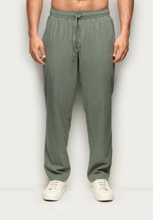 Mężczyzna w luźnych oliwkowozielonych spodniach ze sznurkiem i białych sneakersach, stojący na jednolitym jasnym tle.