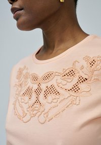Camiseta melocotón con un intrincado diseño bordado y accents de cuentas. Cuello redondo, textura suave y detalle de patrón definido.