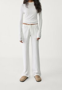 Biały top z długim rękawem i białe spodnie o szerokich nogawkach wykonane z miękkiego materiału, z talią na sznurek i bocznymi kieszeniami. Beżowe wsuwane buty.