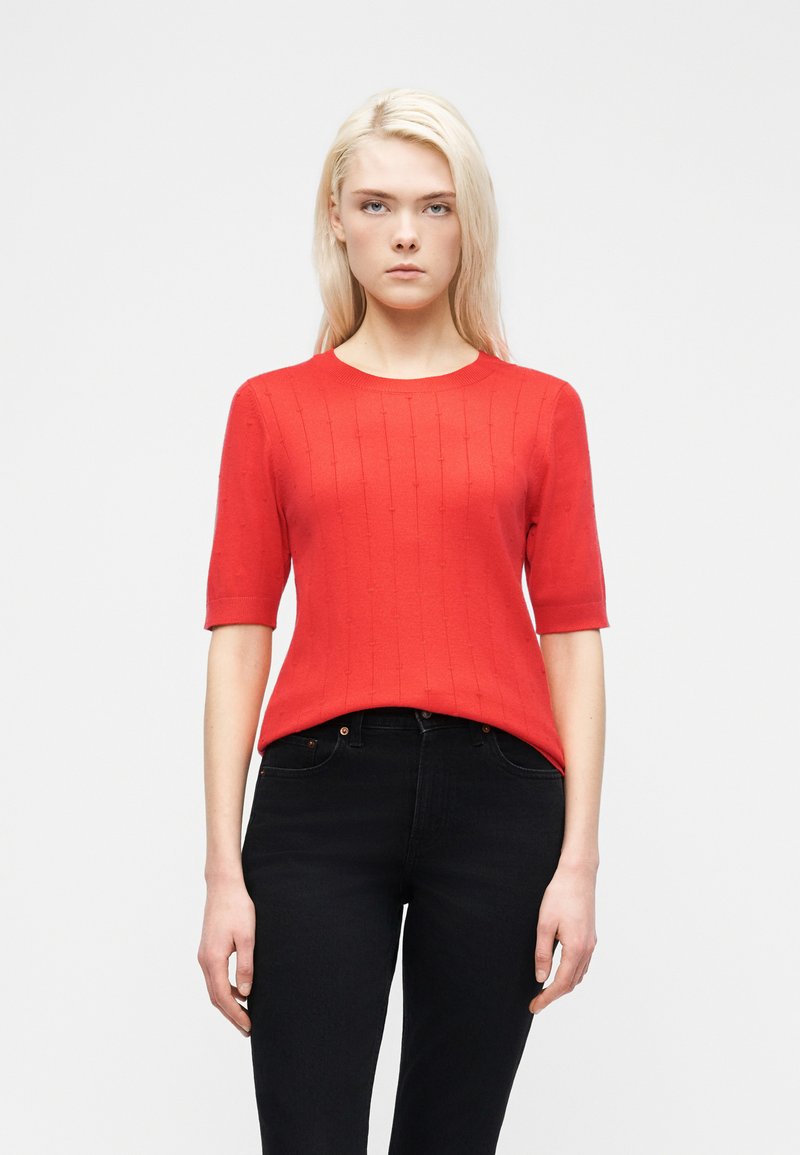 Femme blonde portant un haut en maille rouge à manches courtes et un jean noir taille haute, debout devant un fond blanc uni.