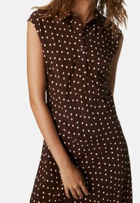 Vestito senza maniche marrone con pois rosa chiaro, con bottoni davanti e colletto, indossato da una donna con capelli lunghi fino alle spalle.