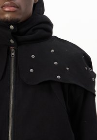 Chaqueta negra con capucha que presenta un cierre frontal, un diseño de hombros único con tachuelas plateadas, una textura suave y una tela negra estructurada.