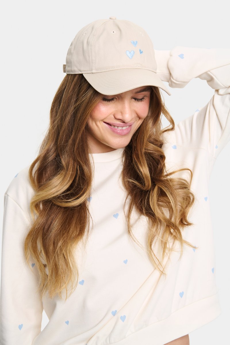 Bézs színű sapka kék szív hímzéssel, puha anyagból készült. A sapkán ívelt napellenző és hátul állítható pánt található.