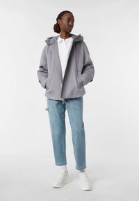 Woman wearing a light gray hooded jacket, white button-up shirt, light blue jeans, and white sneakers standing against a plain background.
