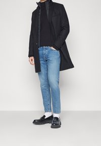 Un abrigo negro largo sobre un suéter negro, combinado con jeans azul claro y zapatos negros de tipo slip-on. Calcetines blancos visibles por encima de los zapatos.