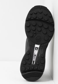 Semelle de basket noire avec des motifs circulaires antidérapants, des rainures surélevées au talon et à l’avant-pied, et une bande métallique portant l’inscription "MICHAEL KORS" à la voûte plantaire.