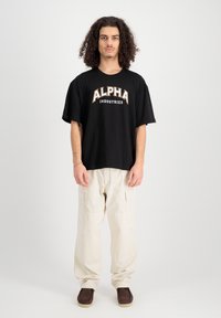 Fekete, oversized póló fehér és narancssárga merész betűkkel az "ALPHA INDUSTRIES" felirattal, világos bézs cargo nadrággal és barna cipőkkel párosítva.