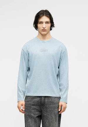 Junger Mann mit schulterlangem dunklem Haar, der ein hellblaues Langarmhemd und ausgewaschene schwarze Jeans trägt und vor einem schlichten Hintergrund steht.