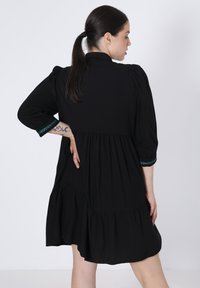 Zwarte jurk met een gelaagd ontwerp, pofmouwen en een cinched taille. De manchetten hebben een turkooise geometrisch patroon. Zachte stoftextuur.