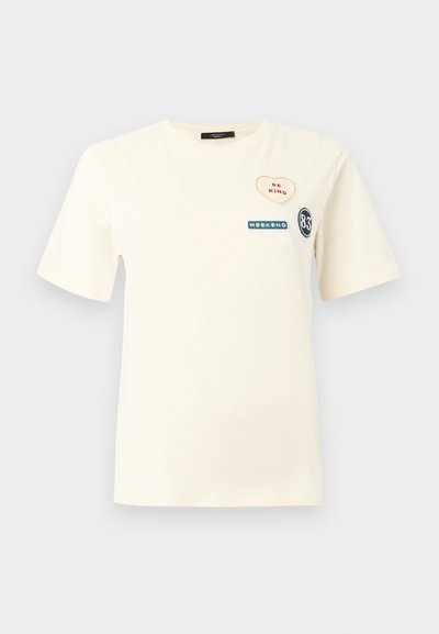 Κοντομάνικο μπλουζάκι σε κρεμ χρώμα με τρία μπαλώματα στο στήθος: καρδιά με τη φράση "ΝΑ ΕΙΣΑΙ ΕΥΓΕΝΙΚΟΣ", ορθογώνιο με τη λέξη "ΣΑΒΒΑΤΟΚΥΡΙΑΚΟ" και κύκλος με τον αριθμό "83".