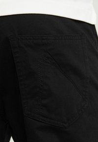 Los pantalones de algodón negros presentan un bolsillo trasero angulado y distintivo con costuras en contraste. La textura suave y la forma ajustada mejoran la funcionalidad.