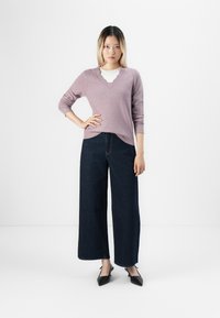 Mulher em pé contra um fundo branco, vestindo um suéter V-decorado em lilás claro, jeans largos escuros e sapatos de salto pretos.