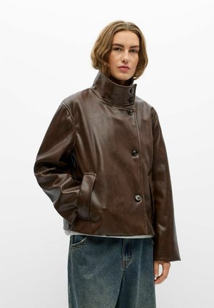 Jeune personne aux cheveux mi-longs portant une veste en cuir marron foncé à col montant et un jean bleu ample sur fond blanc.