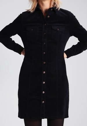 Femme portant une robe noire à manches longues boutonnée avec poches poitrine, debout les mains sur les hanches devant un fond clair uni.
