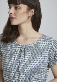 Kurzärmelige Bluse mit rundem Ausschnitt, die ein blaues und grünes geometrisches Muster aufweist, aus leichtem Stoff besteht und eine Raffungsdetail am Kragen hat.