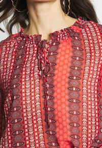 Rote gemusterte Bluse mit gerafftem Ausschnitt und Bändern. Sie verfügt über aufwendige Blumen- und Paisleymuster und besteht aus einem weichen, leichten Stoff.