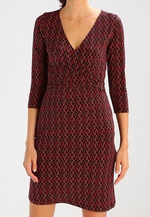 Vrouw die een nauwsluitende jurk tot op de knie draagt met driekwart mouwen en een rood-zwart abstract patroon, met een V-halslijn.