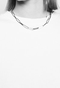 Collier en chaîne argentée avec des maillons rectangulaires, porté par-dessus un haut blanc à col rond, cheveux partiellement visibles et boucle d'oreille pendante d'un côté.