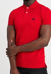 Camisa polo roja de algodón, con cuello abotonado, mangas cortas y un pequeño logo negro encima del dobladillo. Tejido texturizado con aberturas laterales.