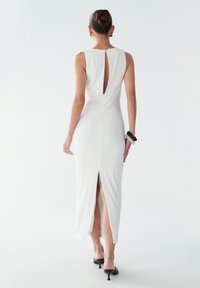 Robe midi blanche texturée avec un dos en trou de serrure, une fente sur le côté et sans manches. Portée avec des sandales à talons noires et un bracelet.