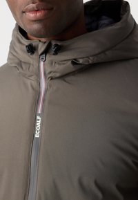 Dark olive green hooded jacket featuring a zipped front, adjustable cords, and contrasting red and white details on the zipper.
