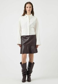 Witte oversized blouse met lange mouwen, twee borstzakken, gecombineerd met een donkerbruine leren uitlopende rok en slouchy bruine kniehoge laarzen.