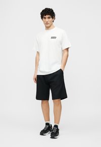 Νέος άνδρας με μπούκλες, φορούντας λευκό T-shirt με σχέδιο, μαύρο σορτς, λευκές κάλτσες και μαύρα αθλητικά παπούτσια, στέκεται ουδέτερα σε γκρι φόντο.