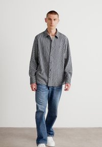 Ριγέ πουκάμισο σε μαύρο και λευκό, συνδυασμένο με μπλε τζιν και λευκά αθλητικά παπούτσια, σε φωτεινό ουδέτερο φόντο.