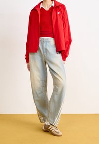 Czerwona lekka kurtka z białym logo, czerwony sweter z prążkowanym wzorem, luźne jasnoniebieskie dżinsy z detalami szwów oraz brązowe sneakersy z białymi paskami.