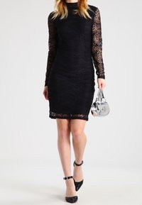 Svart spetsklänning med långa ärmar, figurnära silhuett, blommönster och rundad halsringning. Matchad med blanka svarta klackar och en silverhandväska.