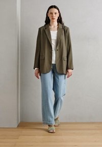 Blazer de gran tamaño en verde oliva con dos bolsillos frontales, top ajustado blanco, vaqueros de pierna ancha en azul claro y sandalias de tiras beige. Textura suave.