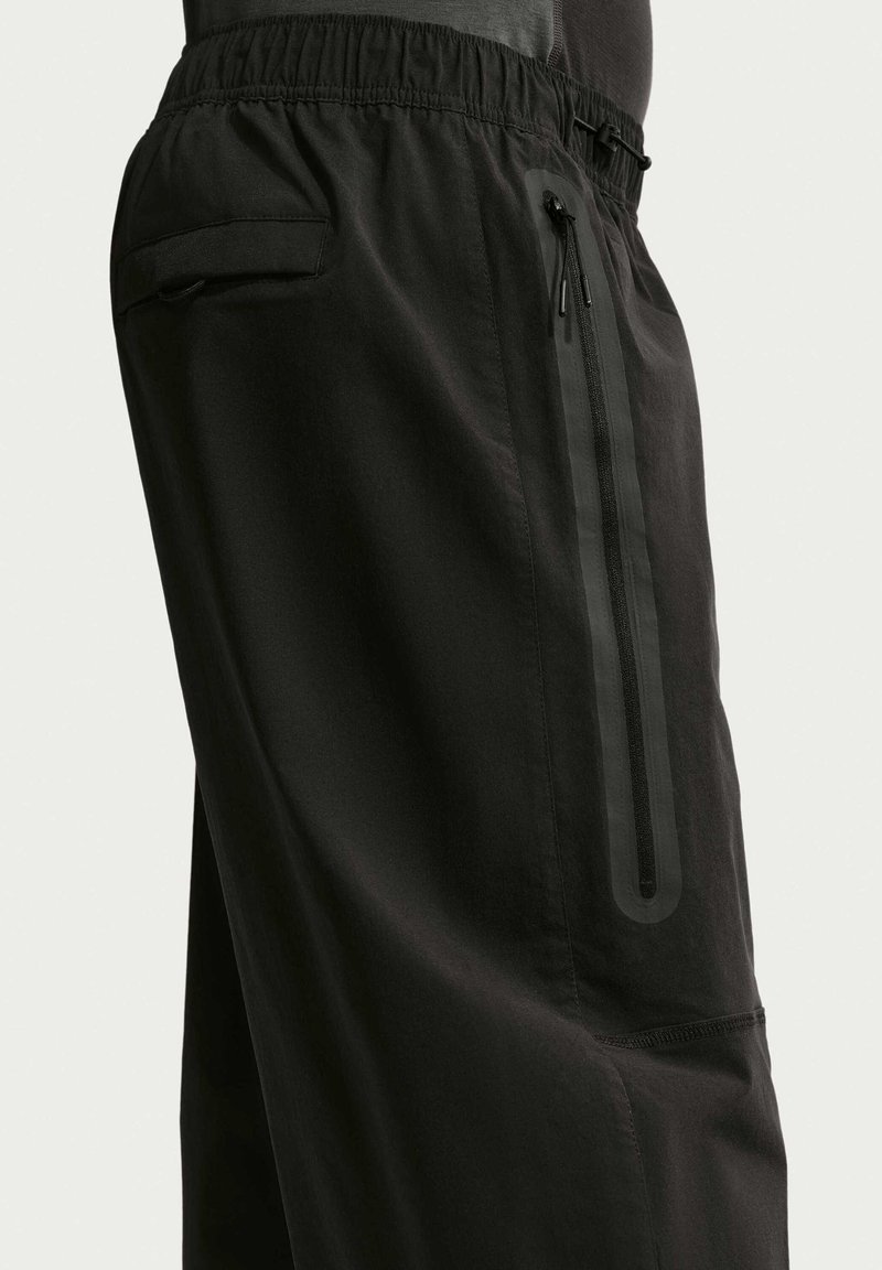 Pantaloni neri con cinturino elastico in vita, dotati di una tasca verticale con zip e una tasca con patta sul fianco laterale.