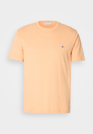Short-sleeve t-shirt in light peach cotton, featuring a small blue logo on the chest. Round neckline with a smooth texture.