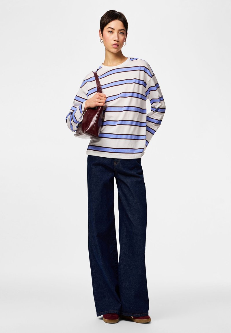 Femme aux cheveux courts et foncés, portant un chemisier à manches longues rayé bleu et blanc, un jean large foncé, des chaussures bordeaux, et tenant un sac à bandoulière bordeaux.