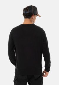 T-shirt noir à manches longues avec un col rond, présentant une texture lisse. Le modèle a des cheveux bouclés et porte une casquette rayée noire avec un accent orange.