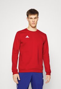 Sweatshirt rouge à manches longues en tissu doux, avec un col rond et un logo Adidas blanc sur la poitrine gauche. Design simple, sans motifs.