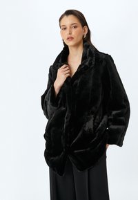 Abrigo de piel sintética negro con un amplio cuello, corte oversize y textura mullida. Cuenta con bolsillos laterales y un dobladillo ligeramente curvado.