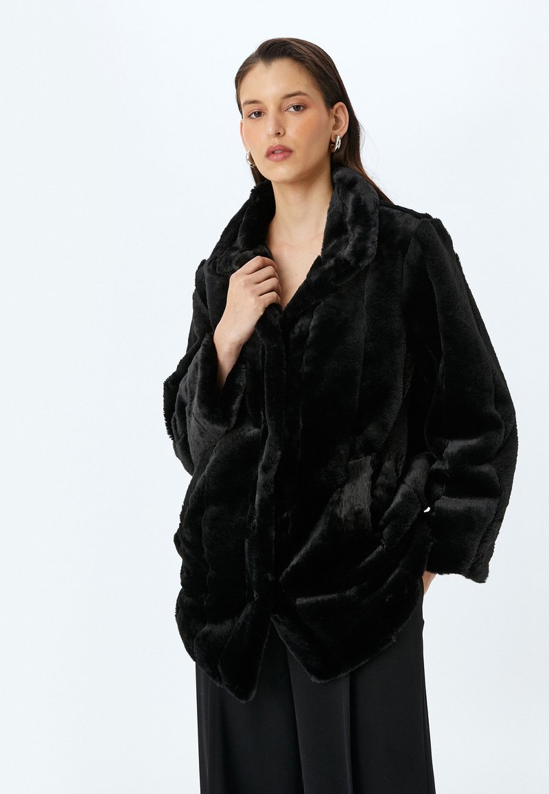 Abrigo de piel sintética negro con un amplio cuello, corte oversize y textura mullida. Cuenta con bolsillos laterales y un dobladillo ligeramente curvado.