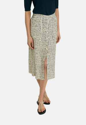 Mujer con falda midi beige con puntos geométricos negros y abertura frontal, combinada con una blusa negra y sandalias beige.